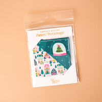 Sugarplum Lane Corner Keeper Fabric Bookmark featuring a pattern of colorful houses and a snow globe, packaged in a clear plastic sleeve.