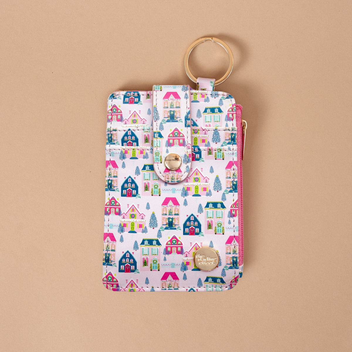 The Sugarplum Lane Keychain Wallet is a vertical zippered pouch featuring a pattern of colorful illustrated houses and Christmas trees, with a snap-button strap and a gold-toned metal key ring.