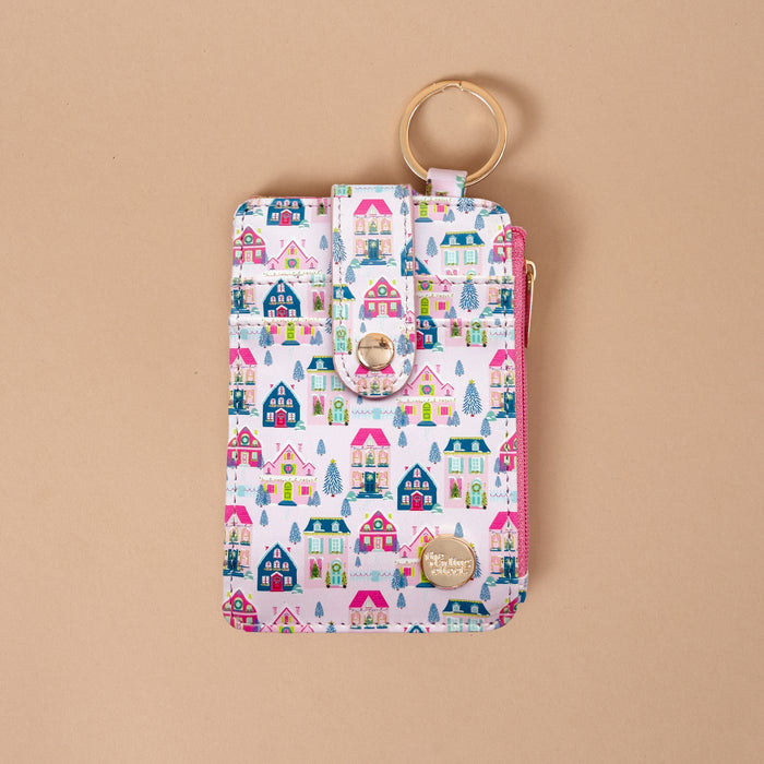 The Sugarplum Lane Keychain Wallet is a vertical zippered pouch featuring a pattern of colorful illustrated houses and Christmas trees, with a snap-button strap and a gold-toned metal key ring.
