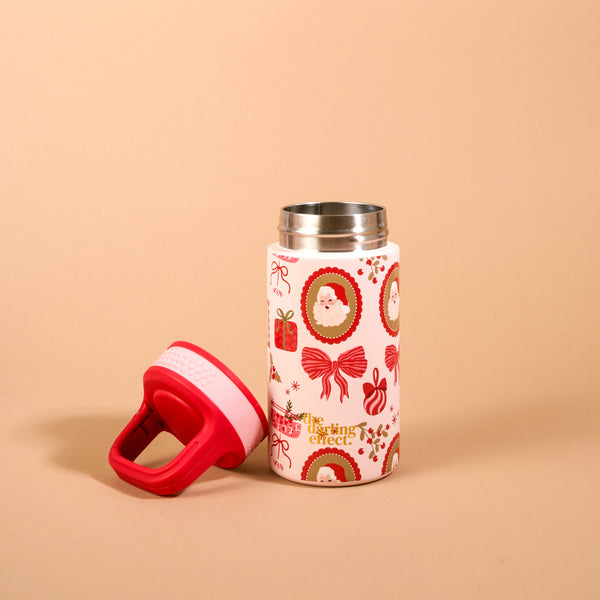 A petite, insulated Mistletoe Magic 6 oz Lil’ Bitty Bottle patterned with Christmas-themed illustrations, including Santa Claus, bows, and gift boxes, with its red screw-top lid sitting beside it.