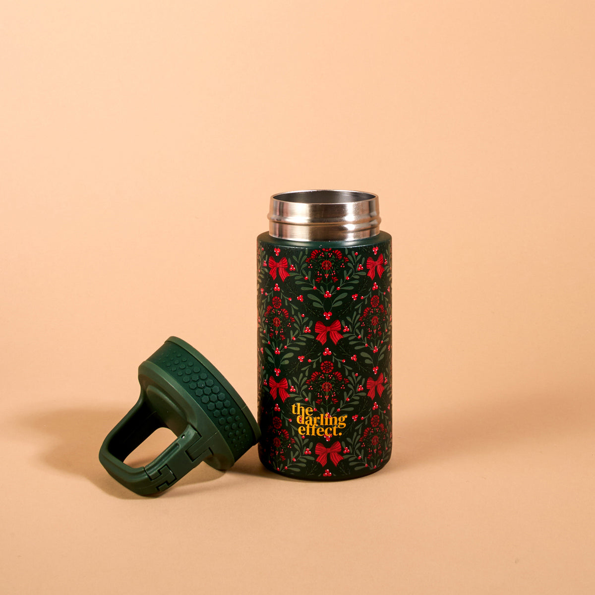 The Darling Effect A Merry Moment 6 oz Lil’ Bitty Bottle is a dark green insulated bottle with a holiday-themed pattern of red bows and holly, shown next to its detached green lid.
