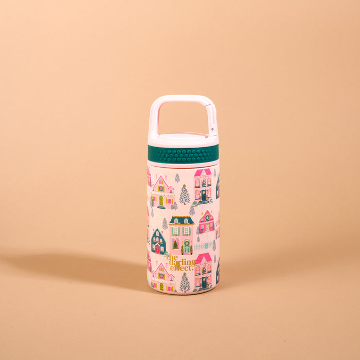The Darling Effect Sugarplum Lane 6 oz Lil’ Bitty Bottle is a petite insulated white water bottle featuring a teal silicone grip, a white carry loop, and a repeating pattern of colorful holiday-themed houses and evergreen trees.