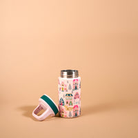 A Sugarplum Lane 6 oz Lil’ Bitty Bottle decorated with a pattern of colorful illustrated houses and evergreen trees, with its matching pink and dark green lid resting beside it.