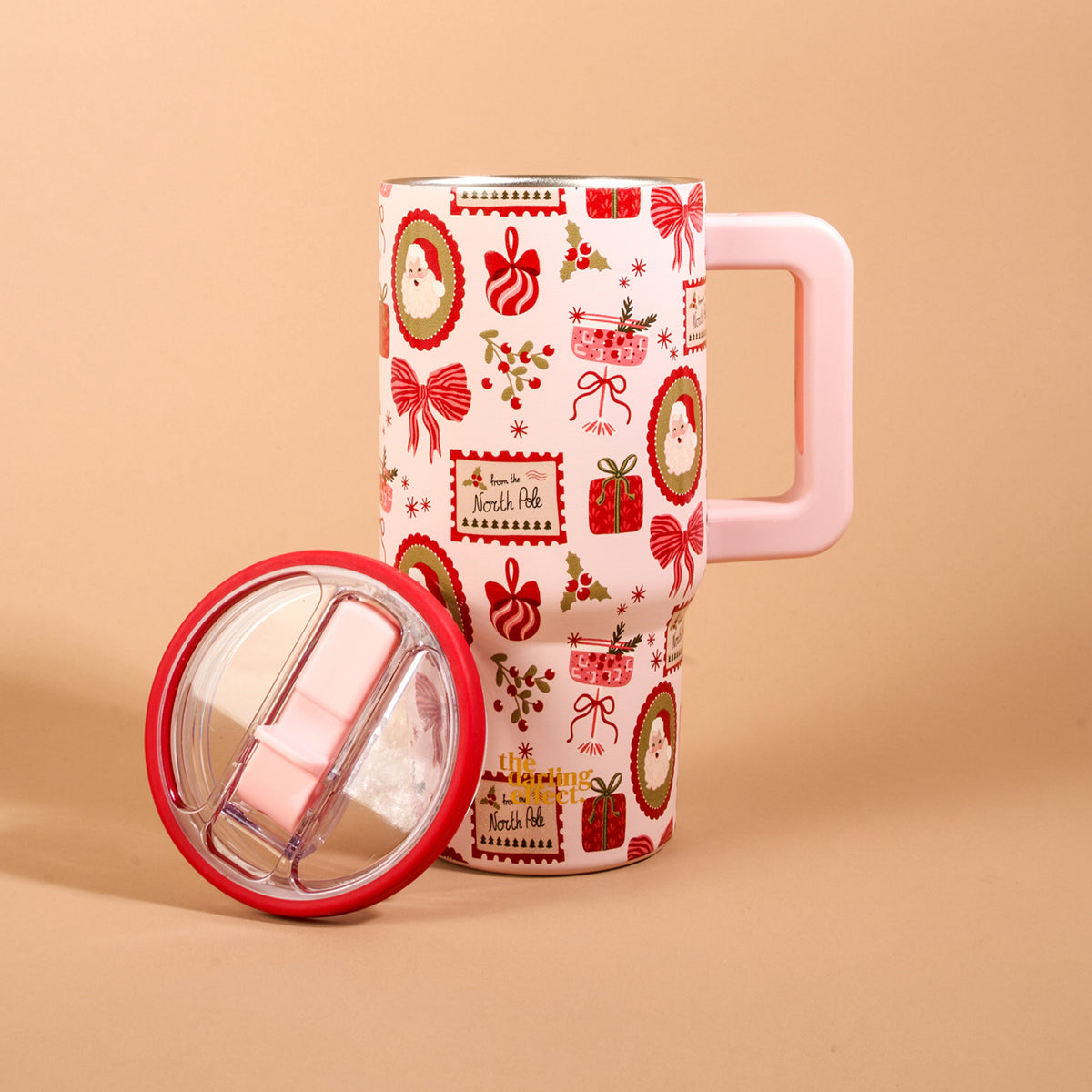 Light pink Mistletoe Magic 20 oz Lifestyle Flip Straw Tumbler with a handle and a pattern of Christmas icons, including Santa, gifts, and bows, with its removable flip straw lid placed in front.