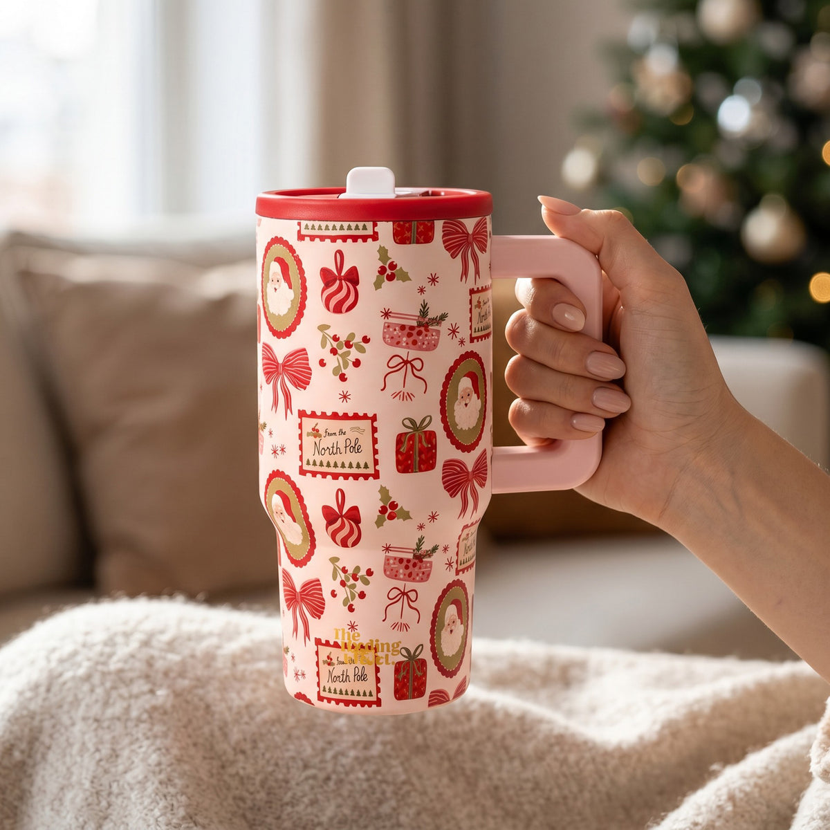 A person holds a tall, light pink 20 oz Mistletoe Magic 20 oz Lifestyle Flip Straw Tumbler patterned with Christmas icons including Santa Claus, gifts, bows, and holly.