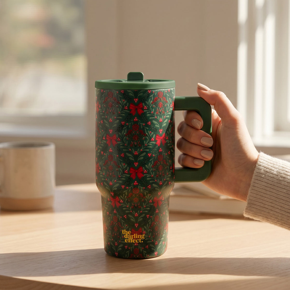 A hand holds a green A Merry Moment 20 oz Lifestyle Flip Straw Tumbler with a handle, featuring a repeating pattern of red bows and holly leaves.