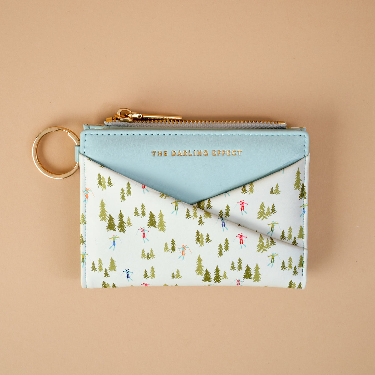 Aprés Ski Luxe Pocket Wallet in light blue vegan leather, featuring a gold zipper and a pocket printed with green evergreen trees and tiny figures skiing.