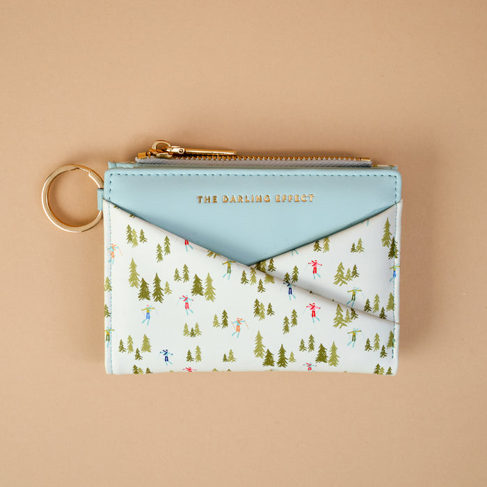 Aprés Ski Luxe Pocket Wallet in light blue vegan leather, featuring a gold zipper and a pocket printed with green evergreen trees and tiny figures skiing.