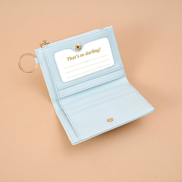 Open light blue vegan leather Aprés Ski Luxe Pocket Wallet with a criss-cross design, featuring a gold key ring and an ID window containing a card that reads Thats so darling! above blank lines.