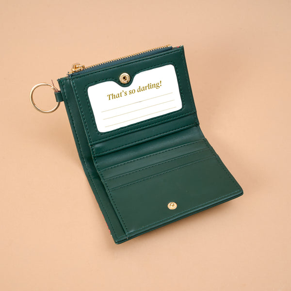 A Merry Moment Luxe Pocket Wallet in dark green vegan leather featuring a criss-cross design, a clear ID window labeled Thats so darling!, and a gold keyring attached to the zipper.