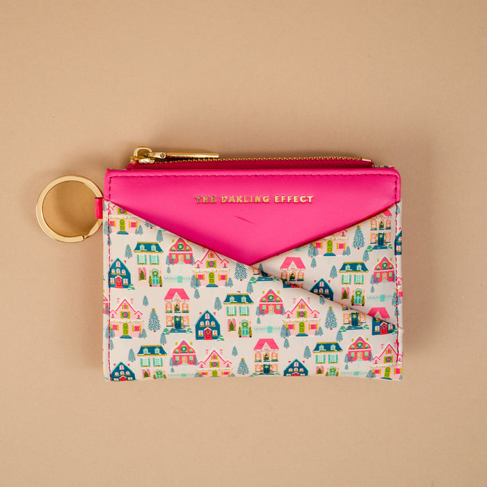 The Sugarplum Lane Luxe Pocket Wallet by The Darling Effect is a hot pink vegan leather pocket wallet with a gold key ring, featuring an envelope-style flap and a repeating pattern of colorful houses and evergreen trees.