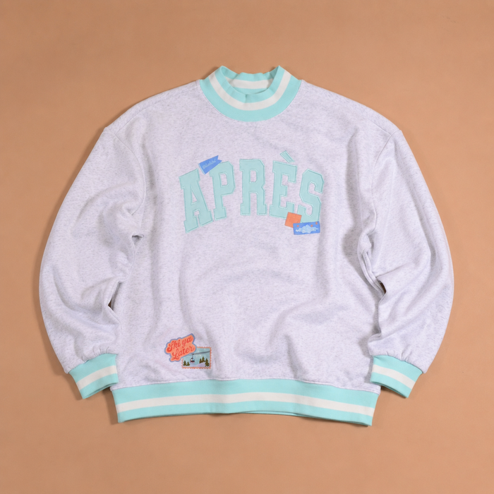 Aprés Ski Mockneck Sweatshirt by The Darling Effect: a light grey heathered mockneck with a relaxed silhouette, turquoise striped trim, and turquoise APRÈS varsity lettering featuring colorful retro-style patches.