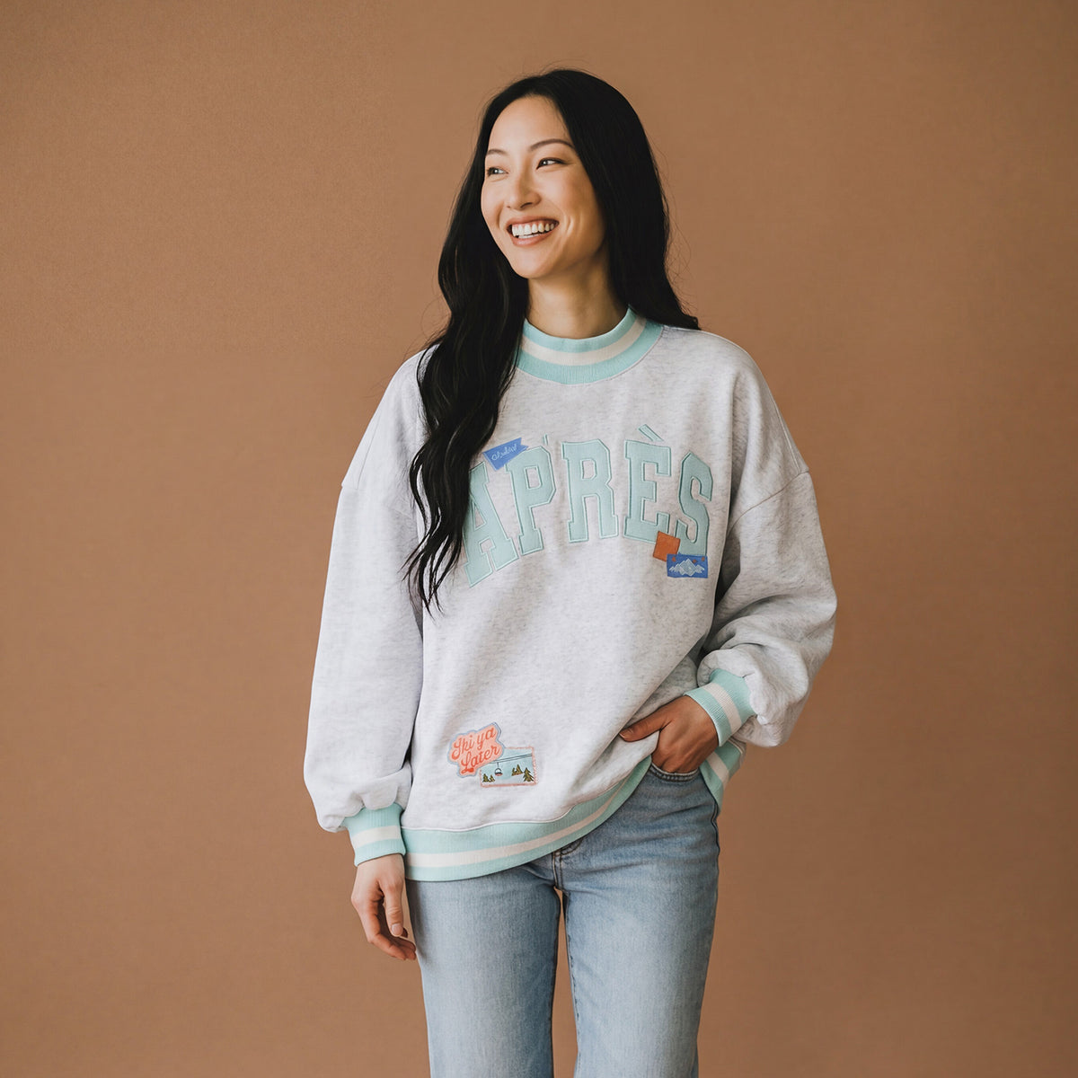 A person in a relaxed-fit Aprés Ski Mockneck Sweatshirt featuring APRÈS lettering, winter-themed patches, and blue and white striped trim.