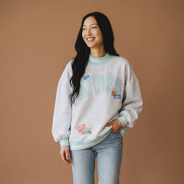 A person in a relaxed-fit Aprés Ski Mockneck Sweatshirt featuring APRÈS lettering, winter-themed patches, and blue and white striped trim.