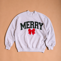 The Darling Effect Merry Bow Mockneck Sweatshirt is a light gray, relaxed-fit mockneck featuring the word MERRY in dark green varsity letters above a red bow graphic.