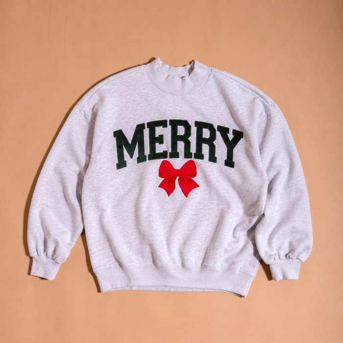 The Darling Effect Merry Bow Mockneck Sweatshirt is a light gray, relaxed-fit mockneck featuring the word MERRY in dark green varsity letters above a red bow graphic.