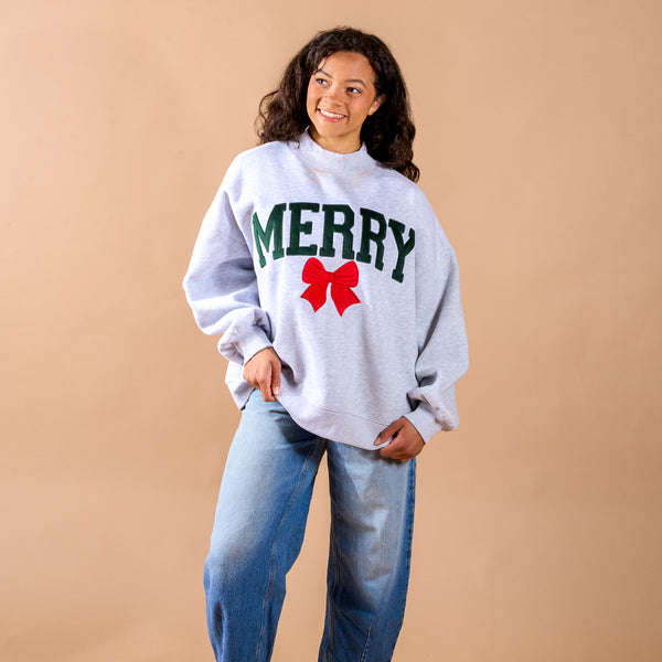 A person wears The Darling Effect Merry Bow Mockneck Sweatshirt, a grey mockneck with a relaxed silhouette featuring green MERRY text and a red bow graphic, paired with blue jeans.