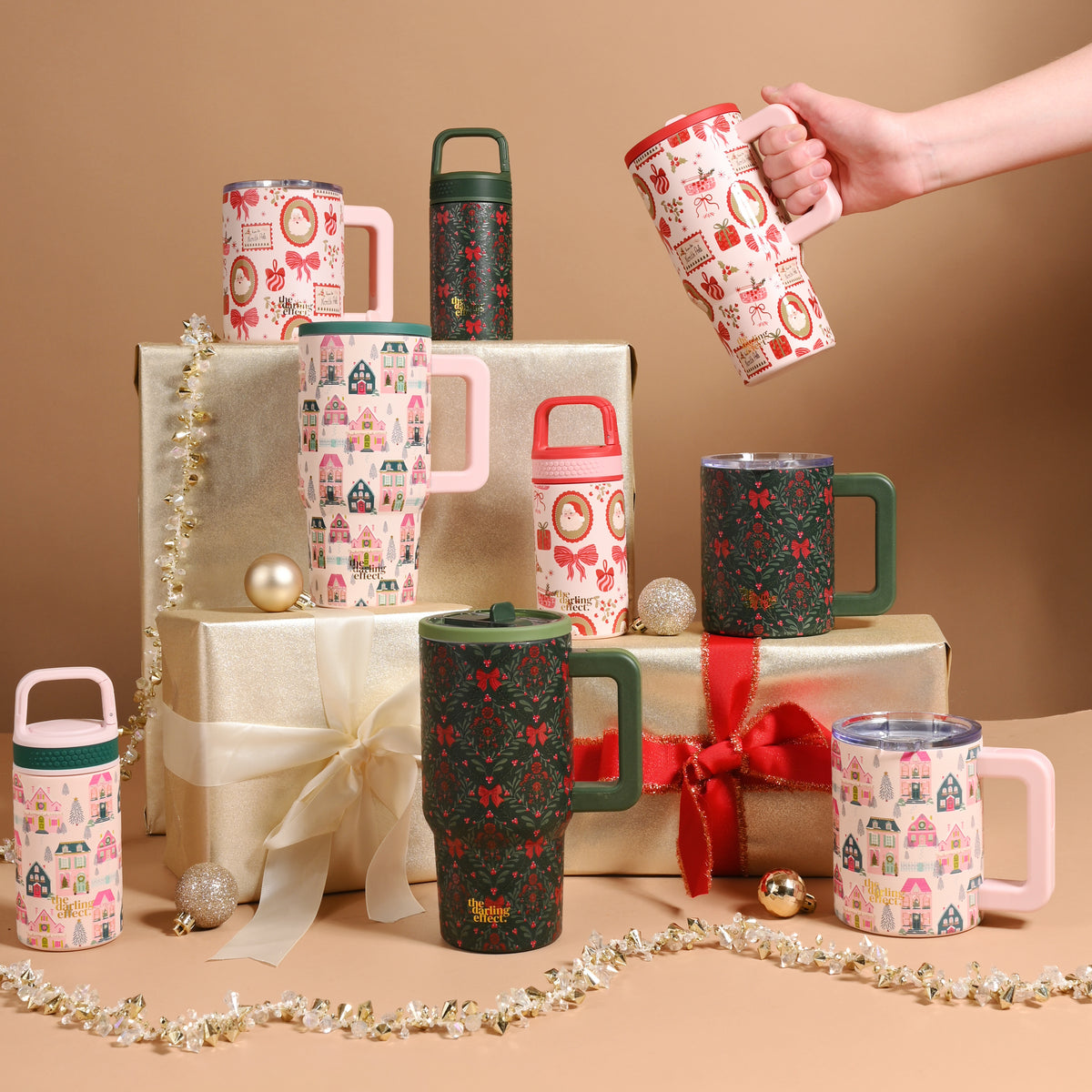 A hand holds up a Sugarplum Lane 14 oz Insulated Mug decorated with holiday motifs above a display of various holiday-themed mugs arranged on gold gift boxes.