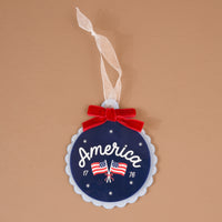 America Acrylic Ornament: A round blue acrylic ornament featuring the word America, two American flags, the years 17 and 76, and a red velvet bow with a white ribbon hanger.