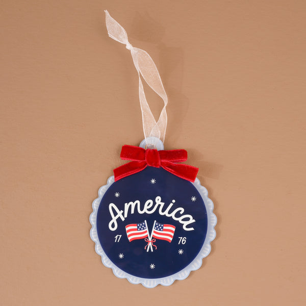 America Acrylic Ornament: A round blue acrylic ornament featuring the word America, two American flags, the years 17 and 76, and a red velvet bow with a white ribbon hanger.