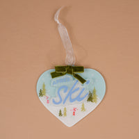Aprés Ski Acrylic Ornament featuring a heart-shaped design with a winter ski scene, blue script, a green velvet bow, and a ribbon hanger.