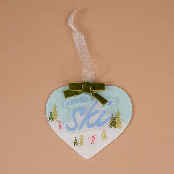 Aprés Ski Acrylic Ornament featuring a heart-shaped design with a winter ski scene, blue script, a green velvet bow, and a ribbon hanger.