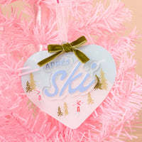 Aprés Ski Acrylic Ornament featuring a heart-shaped acrylic design with a green ribbon bow, text, and illustrations of skiers and pine trees for festive holiday styling.