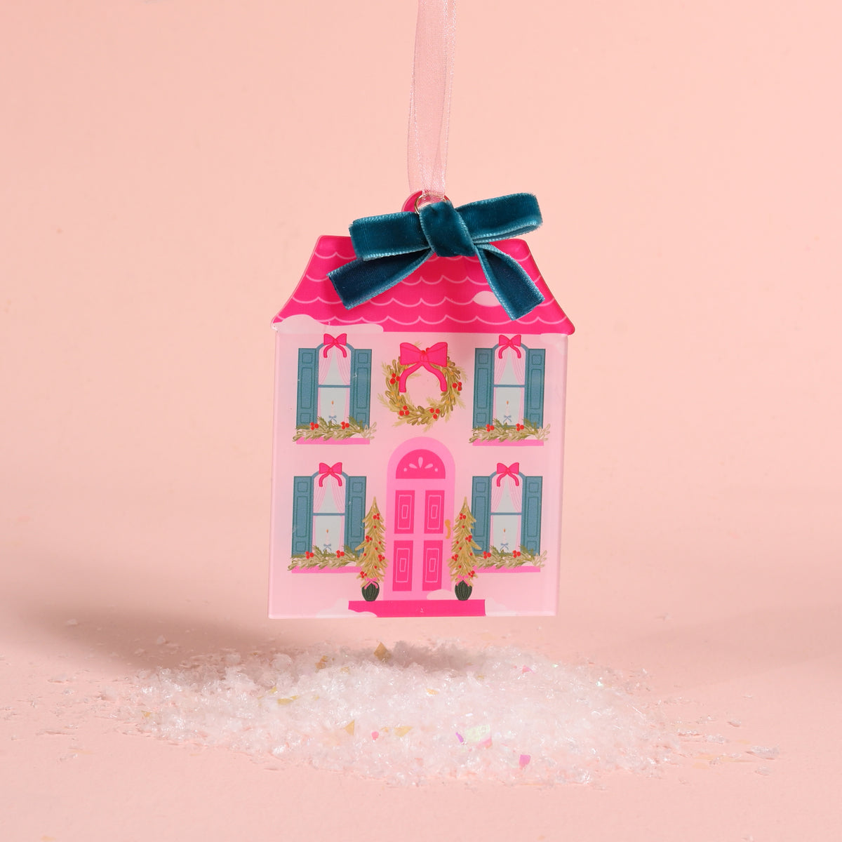 A pink, house-shaped BowBelle Manor Acrylic Ornament by The Darling Effect, decorated with a wreath and blue velvet ribbon bow, hanging above a pile of faux snow.