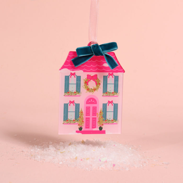 A pink, house-shaped BowBelle Manor Acrylic Ornament by The Darling Effect, decorated with a wreath and blue velvet ribbon bow, hanging above a pile of faux snow.