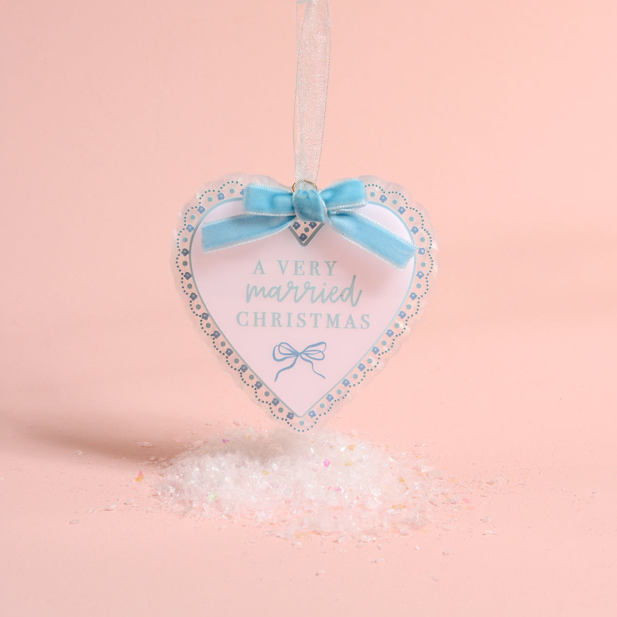A Very Married Christmas 2026 Acrylic Ornament hanging above a pile of faux snow, featuring blue text and a blue ribbon bow at the top for festive holiday styling.