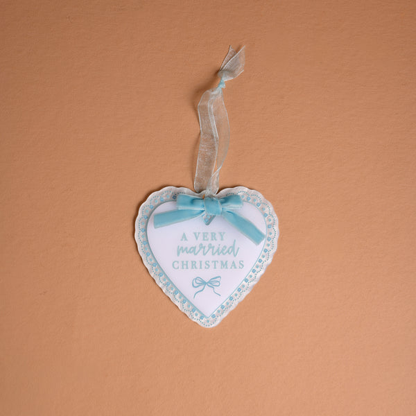 A Very Married Christmas 2026 Acrylic Ornament, featuring a heart-shaped white acrylic design with a blue scalloped border and a light blue ribbon bow.