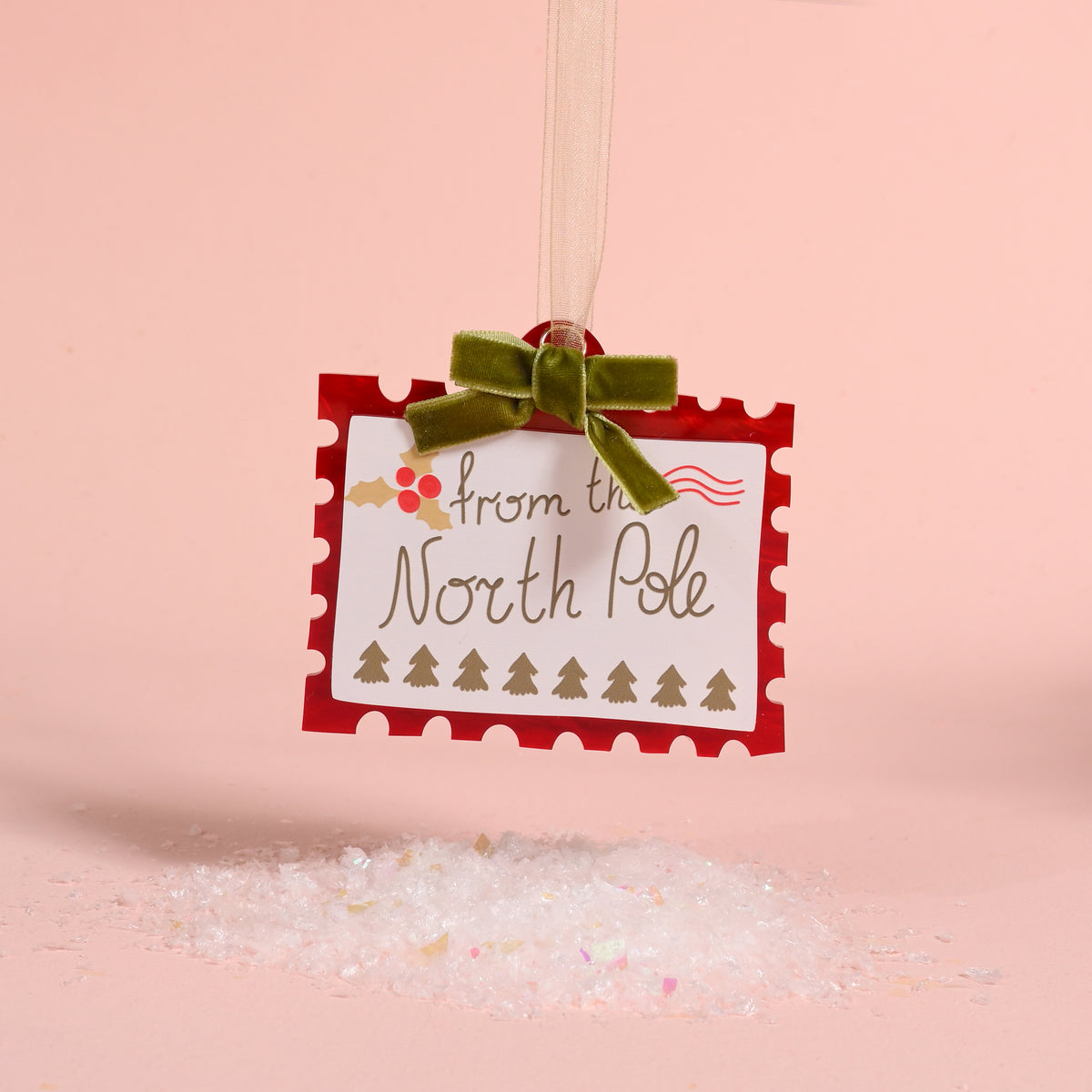 North Pole Stamp Acrylic Ornament shaped like a postage stamp reading from the North Pole, decorated with holly and tiny trees, and finished with a green velvet ribbon for holiday styling.