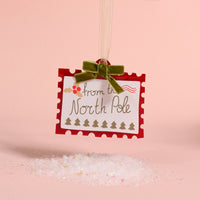 North Pole Stamp Acrylic Ornament shaped like a postage stamp reading from the North Pole, decorated with holly and tiny trees, and finished with a green velvet ribbon for holiday styling.