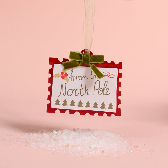 North Pole Stamp Acrylic Ornament shaped like a postage stamp reading from the North Pole, decorated with holly and tiny trees, and finished with a green velvet ribbon for holiday styling.