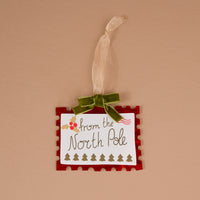 North Pole Stamp Acrylic Ornament shaped like a postage stamp with a red border, holiday North Pole theme, holly, trees, and a green ribbon hanger.