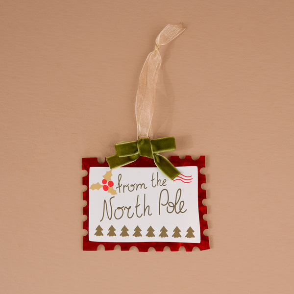 North Pole Stamp Acrylic Ornament shaped like a postage stamp with a red border, holiday North Pole theme, holly, trees, and a green ribbon hanger.