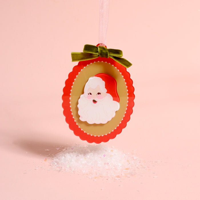 The Darling Effect Retro Santa Acrylic Ornament is an oval-shaped ornament featuring a vintage-style Santa, topped with a green velvet bow and a pink ribbon hanger, displayed over a small pile of faux snow.