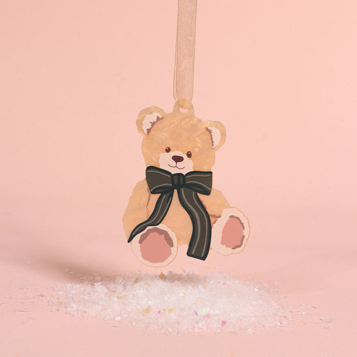 A Teddy Bear Theo Acrylic Ornament featuring a black bow, hanging above a pile of faux snow.