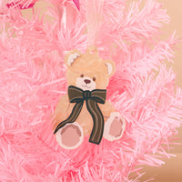 A tan acrylic Teddy Bear Theo Acrylic Ornament with a dark striped bow hangs on a pink artificial tree for festive holiday styling.