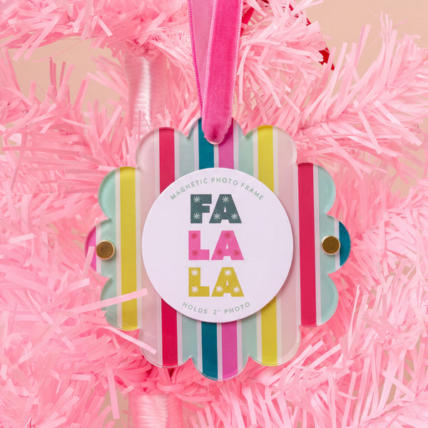 A Fa La La Scalloped Round Picture Frame Ornament hangs on a pink Christmas tree, featuring a scalloped, multi-colored striped round frame with a white circular center that reads FA LA LA and HOLDS 2 PHOTO.