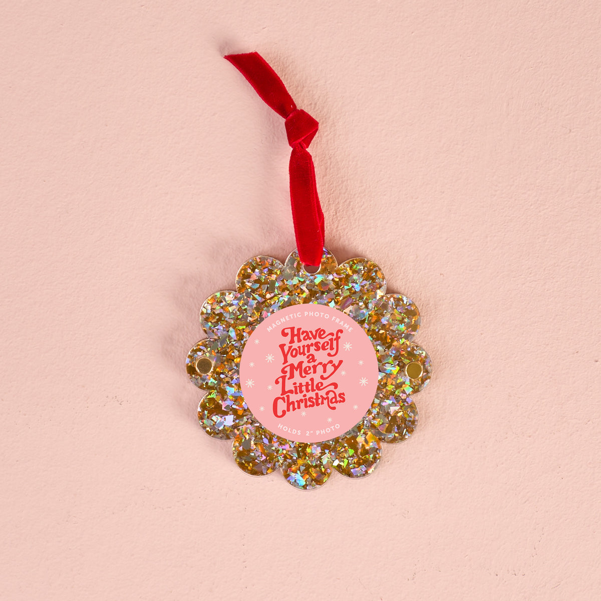 The Darling Effect Gold Glitter Scalloped Round Picture Frame Ornament with a red ribbon hangs on a light pink background. Its pink center displays "Have Yourself a Merry Little Christmas" in white text.