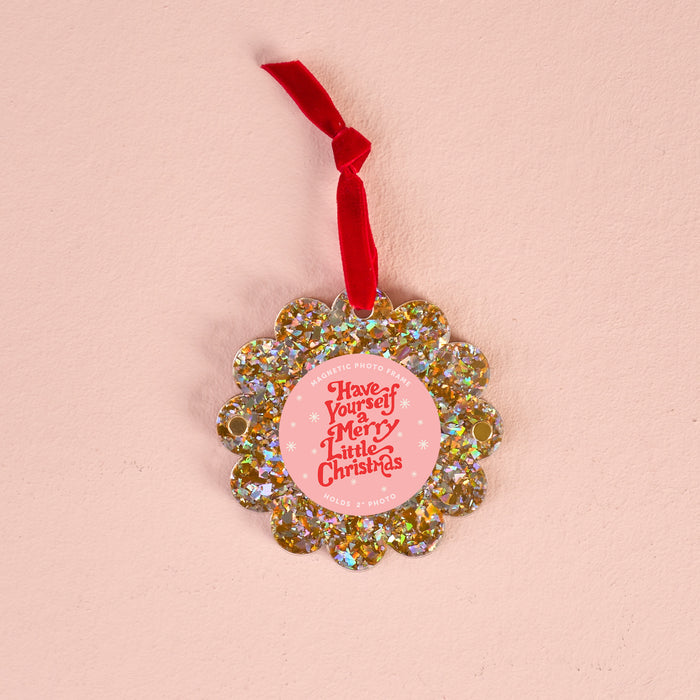 The Darling Effect Gold Glitter Scalloped Round Picture Frame Ornament with a red ribbon hangs on a light pink background. Its pink center displays "Have Yourself a Merry Little Christmas" in white text.