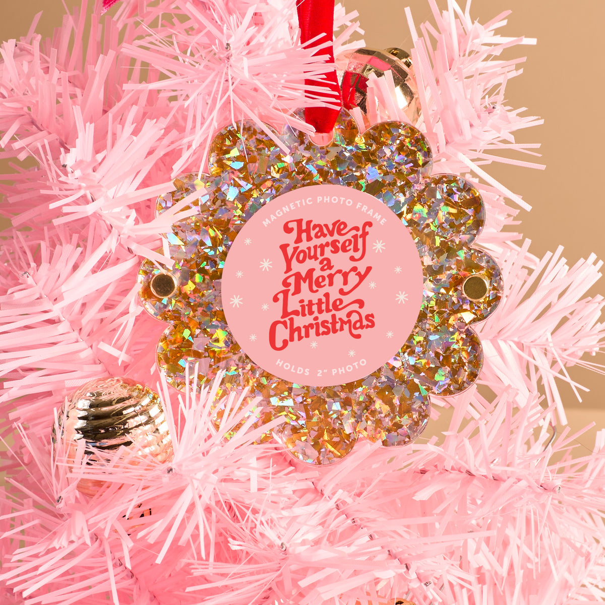 A Gold Glitter Scalloped Round Picture Frame Ornament by The Darling Effect hangs on a light pink Christmas tree, featuring a floral design and the words "Have Yourself a Merry Little Christmas" in red and white. A gold bauble is also visible.