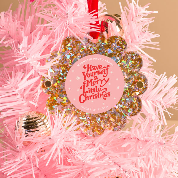 A Gold Glitter Scalloped Round Picture Frame Ornament by The Darling Effect hangs on a light pink Christmas tree, featuring a floral design and the words "Have Yourself a Merry Little Christmas" in red and white. A gold bauble is also visible.