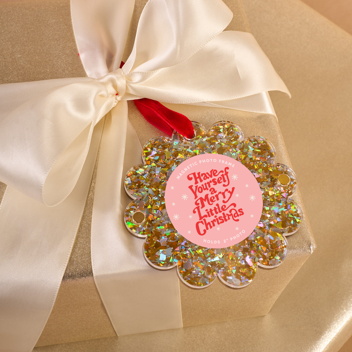 A gold gift box with a white ribbon is topped with The Darling Effect’s Gold Glitter Scalloped Round Picture Frame Ornament, featuring “Have Yourself a Merry Little Christmas” in red on a pink background.