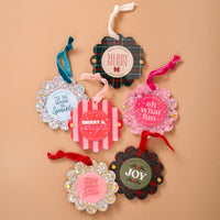 Six festive holiday gift tags with scalloped edges and cheerful sayings, like Merry & bright and JOY, on a tan background—perfect for pairing with The Darling Effect’s Gold Glitter Scalloped Round Picture Frame Ornament.