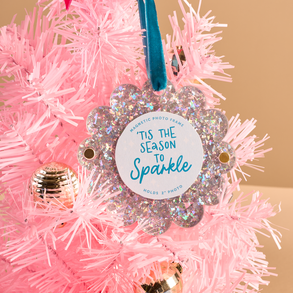The Darling Effect Silver Glitter Scalloped Round Picture Frame Ornament, reading Tis the season to sparkle with a blue velvet ribbon hanger, hanging on a pink artificial Christmas tree.