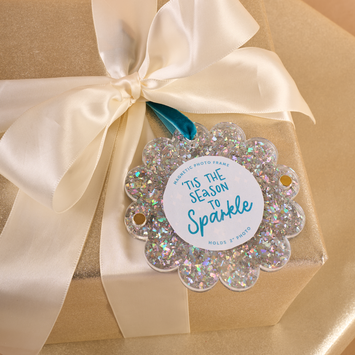A Silver Glitter Scalloped Round Picture Frame Ornament with a center label that reads Tis the Season to Sparkle, resting on a gold gift box with a white ribbon.