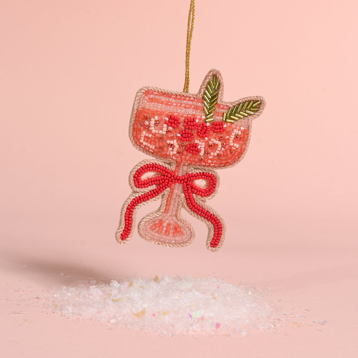 The Darling Effect Cranberry Cosmo handcrafted beaded ornament shaped like a cocktail glass with a garnish and a bow, hanging above a pile of faux snow.