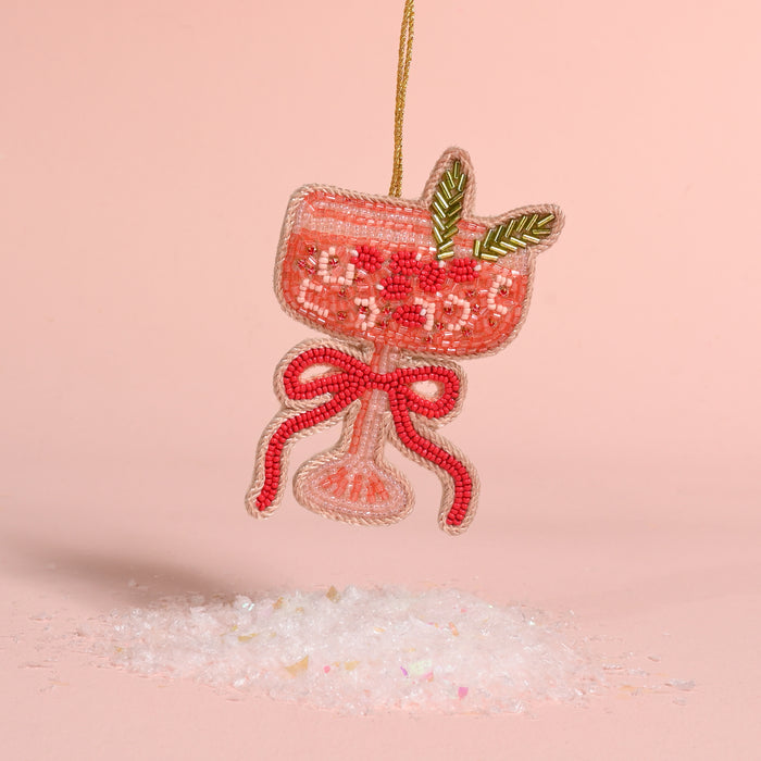 The Darling Effect Cranberry Cosmo handcrafted beaded ornament shaped like a cocktail glass with a garnish and a bow, hanging above a pile of faux snow.